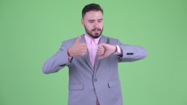 Kafası karışmış genç sakallı işadamı başparmak yukarı ve başparmak aşağı arasında seçim
