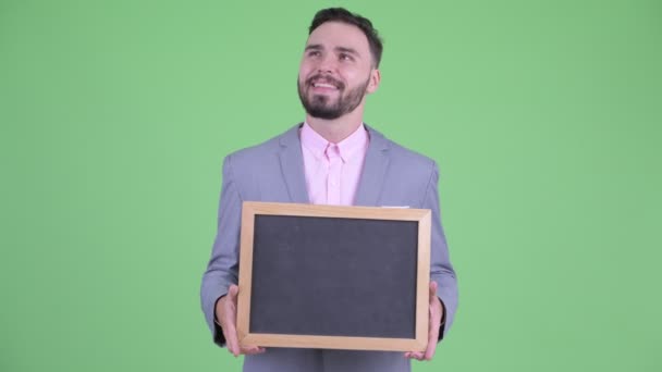 Jovem empresário barbudo feliz pensando enquanto segurando quadro negro — Vídeo de Stock