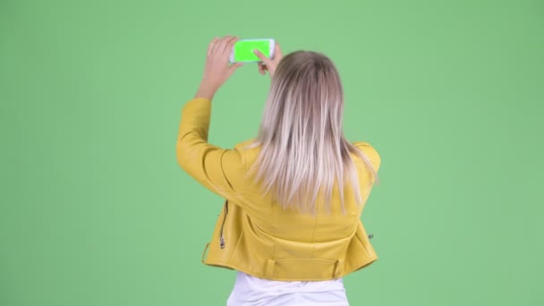 Achteruitkijk van jonge rebelse blonde vrouw die foto met telefoon neemt — Stockvideo