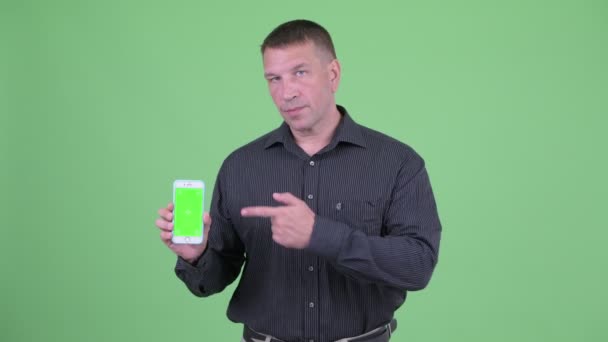 マッチョ成熟したビジネスマンは、携帯電話を示し、親指を与える — ストック動画