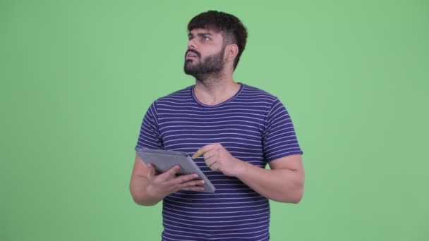 Happy young overweight bearded Indian man thinking while using digital tablet — Stock Video