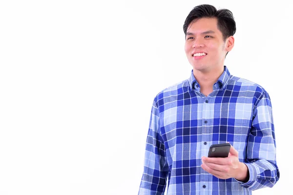 Szczęśliwy młody przystojny Asian Hipster mężczyzna myślący podczas przy pomocy telefon — Zdjęcie stockowe