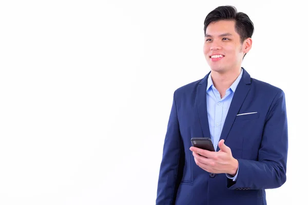 Happy young handsome Asian businessman thinking while using phone — Stock Photo, Image
