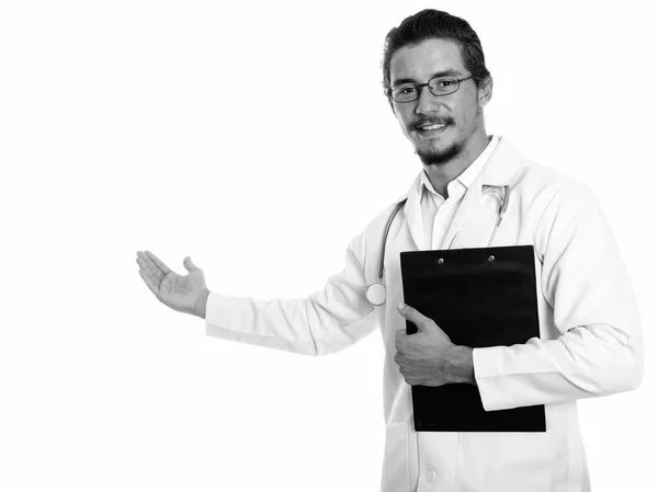 Studio plan de jeune homme heureux médecin souriant tout en montrant quelque chose et tenant presse-papiers isolé sur fond blanc — Photo