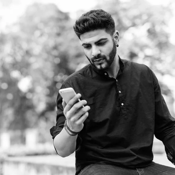 Jeune bel homme indien utilisant un téléphone portable tout en étant assis sur le sol en béton au parc — Photo