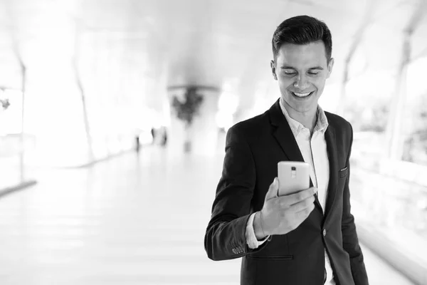 Молодой красивый бизнесмен пользуется телефоном на открытом воздухе, улыбаясь — стоковое фото