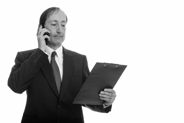 Studioaufnahme eines reifen Geschäftsmannes, der auf dem Handy spricht, während er in der Zwischenablage liest — Stockfoto