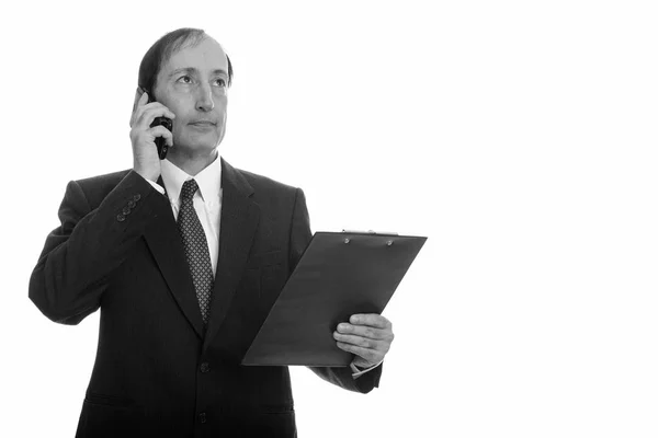 Studio ujęcie przemyślanego dojrzałego biznesmena rozmawiającego przez telefon komórkowy trzymając schowek — Zdjęcie stockowe