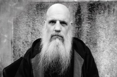 Portrait of mature bald man with long gray beard against grunge concrete wall outdoors in black and white