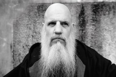 Portrait of mature bald man with long gray beard against grunge concrete wall outdoors in black and white