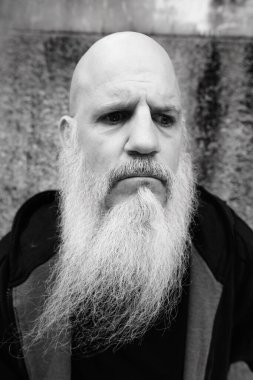 Portrait of mature bald man with long gray beard against grunge concrete wall outdoors in black and white