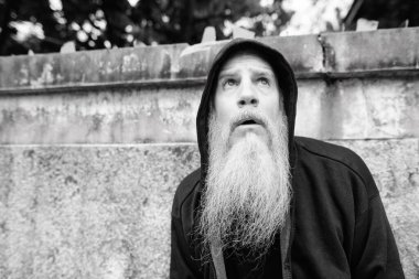 Portrait of mature bald man with long gray beard against grunge concrete wall outdoors in black and white