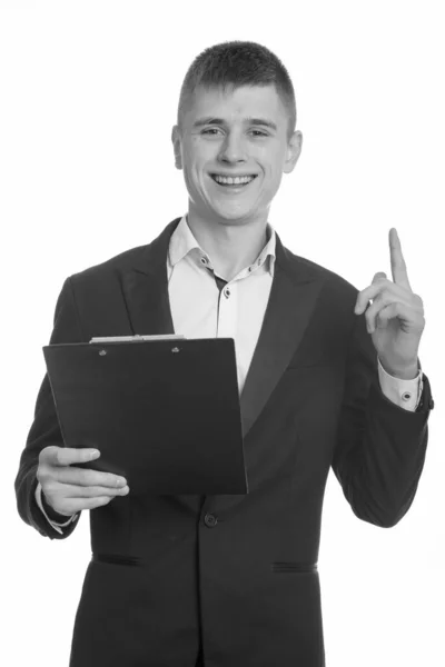 Estúdio tiro de jovem empresário feliz sorrindo enquanto segurando prancheta e apontando o dedo para cima — Fotografia de Stock