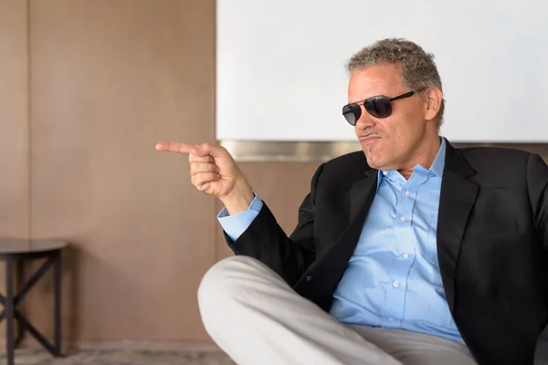 Hombre de negocios hispano maduro con gafas de sol apuntando con el dedo y sentado en la oficina — Foto de Stock