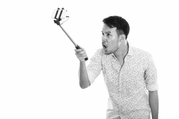 Fiatal dühös ázsiai férfi csinál szelfi kép mobiltelefon, miközben a kezében szelfi bottal — Stock Fotó