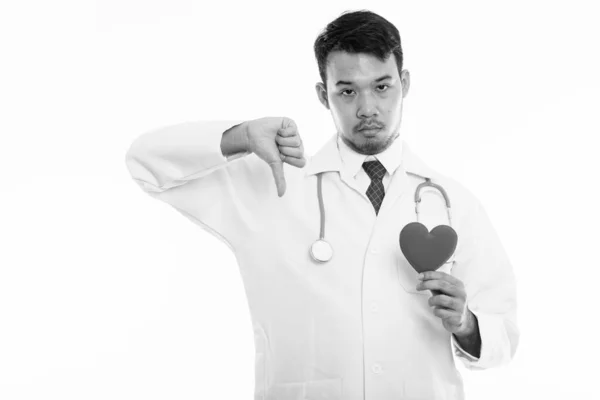 Estudio disparo de joven asiático hombre médico sosteniendo rojo corazón y dando pulgar hacia abajo — Foto de Stock