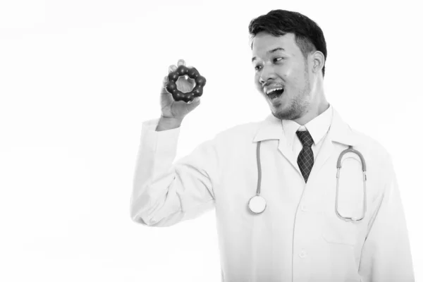 Estudio disparo de joven feliz asiático hombre médico sonriendo mientras sostiene y mirando donut — Foto de Stock