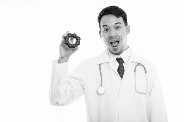 Studio strzał młody szczęśliwy Azji mężczyzna lekarz uśmiechając się i otwierając usta trzymając pączka — Zdjęcie stockowe