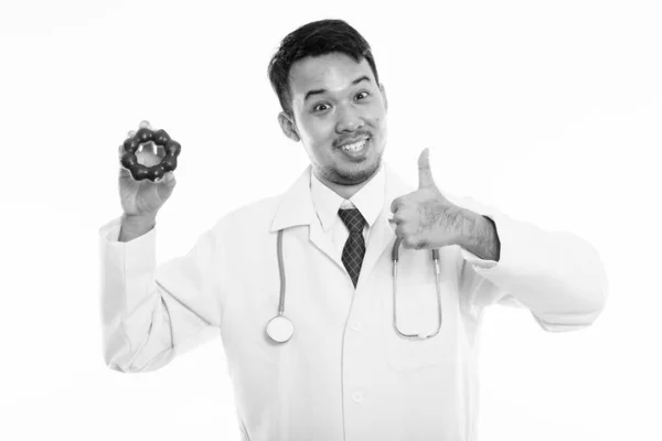 Stúdió lövés fiatal boldog ázsiai férfi orvos mosolyog, miközben a kezében fánk és feladja hüvelykujját — Stock Fotó