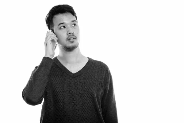 Studio shot of young Asian man talking on mobile phone while thinking — Stock Photo, Image