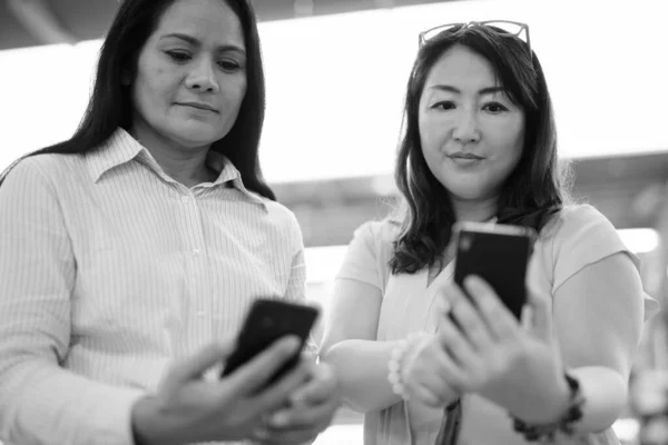 Twee volwassen Aziatische vrouwen samen op het station met behulp van mobiele telefoon — Stockfoto