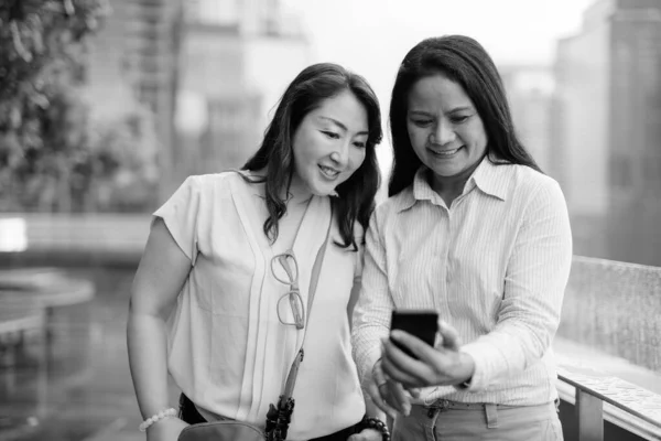 Twee volwassen Aziatische vrouwen samen met behulp van mobiele telefoon in de stad — Stockfoto