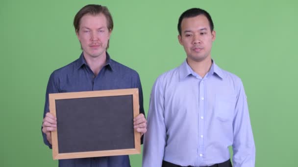 Két stresszes, többnemzetiségű üzletember, akik táblát tartanak és hüvelykujjal mutogatnak. — Stock videók