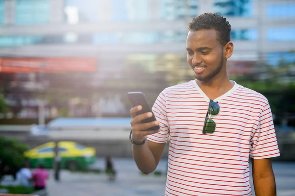 Glad ung afrikansk skäggig man ler när du använder telefon i staden — Stockfoto