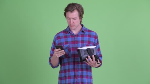 Счастливый молодой хипстер, пользующийся телефоном, держа гарнитуру виртуальной реальности — стоковое видео