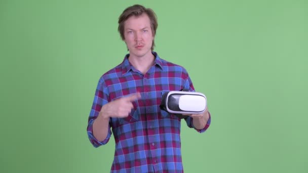 Νεαρός όμορφος hipster άντρας κρατώντας και δείχνοντας τα ακουστικά εικονικής πραγματικότητας — Αρχείο Βίντεο