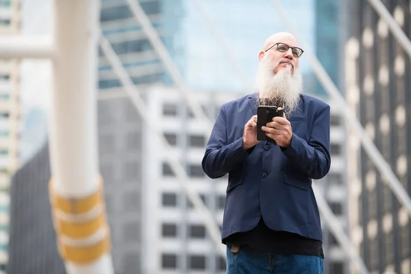 Mature bearded bald businessman thinking while using phone in the city outdoors