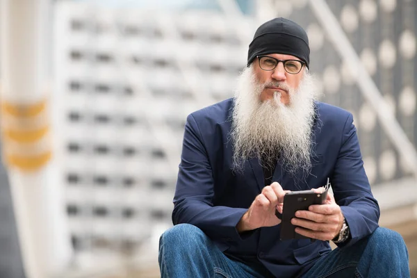 Volwassen bebaarde zakenman zitten terwijl het gebruik van de telefoon in de stad buiten — Stockfoto