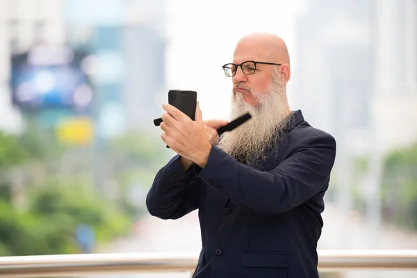 Mature bearded bald businessman brushing beard in the city outdoors