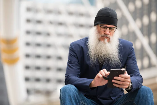 Volwassen bebaarde zakenman zitten terwijl het gebruik van de telefoon in de stad buiten — Stockfoto