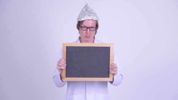 Młody człowiek lekarz z folią aluminiową kapelusz trzymając tablicę i patrząc zszokowany — Wideo stockowe