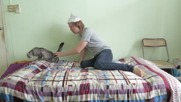Joven asustado usando sombrero de papel de aluminio y entrando en pánico con gato en el dormitorio — Vídeo de stock