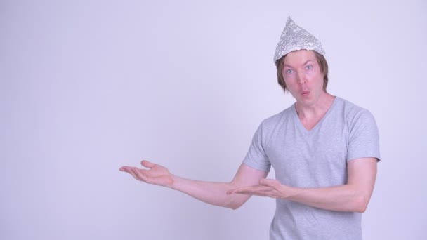 Joven feliz con sombrero de papel de aluminio mostrando algo y mirando sorprendido — Vídeos de Stock