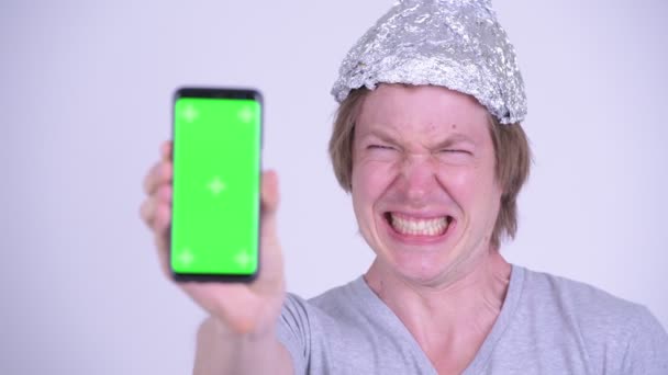 Inför stressad ung man med aluminiumfolie hatt visar telefonen och ser rädd — Stockvideo