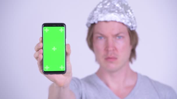Лицо напряженного молодого человека в шляпе из фольги показывает телефон и выглядит испуганным — стоковое видео
