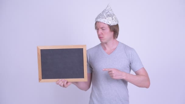 Joven con sombrero de papel de aluminio sosteniendo y señalando la pizarra — Vídeos de Stock