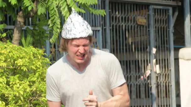 Zestresowany młody człowiek w kapeluszu z folii aluminiowej jako koncepcja teorii spiskowej panikuje na zewnątrz — Wideo stockowe