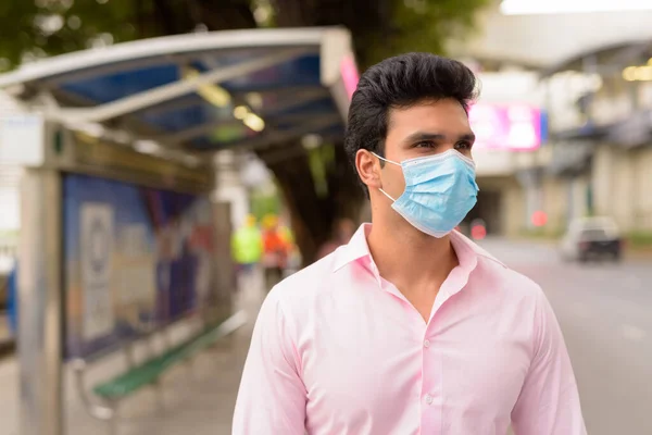 Portrait of young Indian businessman with mask for protection from corona virus outbreak at the bus stop in the city