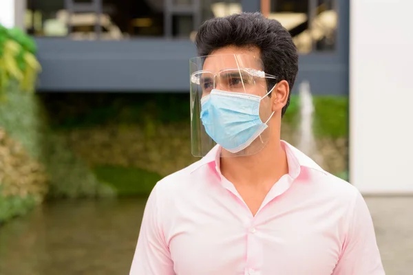 Portrait of young Indian businessman with mask and face shield for protection from corona virus outbreak outside modern building