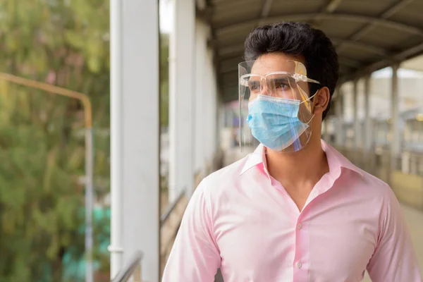 Portrait of young Indian businessman with mask and face shield for protection from corona virus outbreak at footbridge in the city