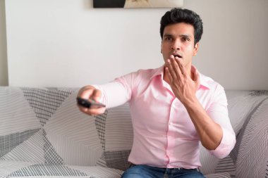Portrait of young handsome Indian businessman sitting on sofa in the living room at home