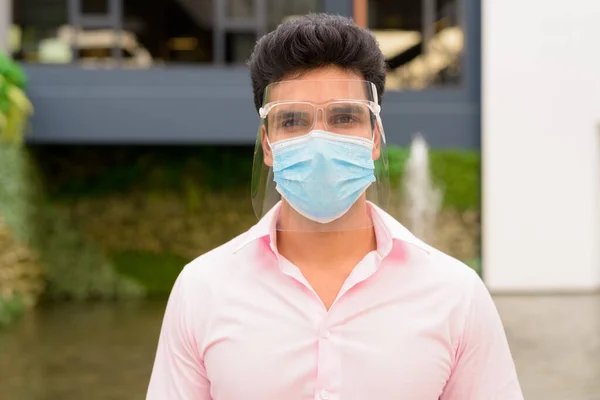 Portrait of young Indian businessman with mask and face shield for protection from corona virus outbreak outside modern building