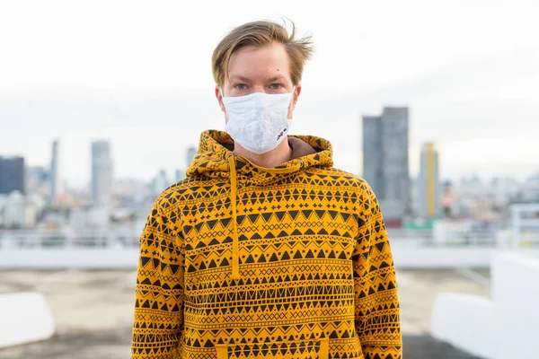 Portrait of young man with mask for protection from corona virus outbreak against view of the city at rooftop of building outdoors