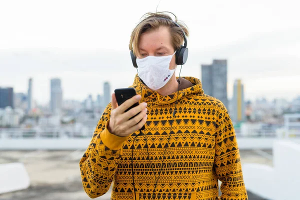 Portrait of young man with mask for protection from corona virus outbreak against view of the city at rooftop of building outdoors