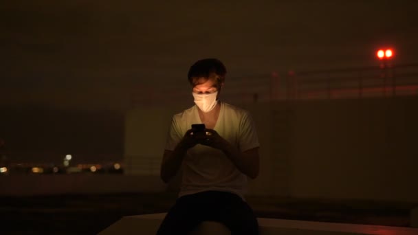 Mladý muž s maskou pomocí telefonu a sedí venku během bouřlivého počasí v noci — Stock video
