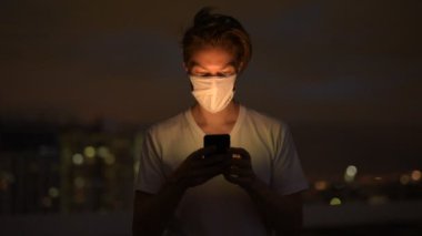 Fırtınalı bir gecede dışarıda telefon kullanan maskeli genç bir adamın yüzü.